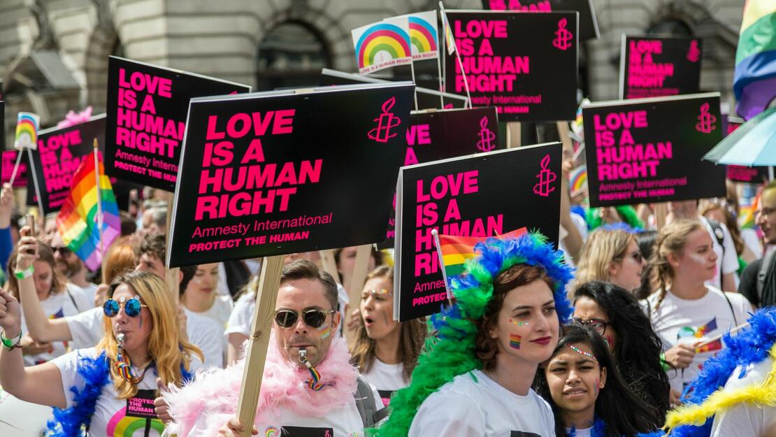 Bilde fra en pride-parade. Flere mennesker med regnbueflagg og plakater fra Amnesty International hvor det står "love is a human right" og "protect the human".