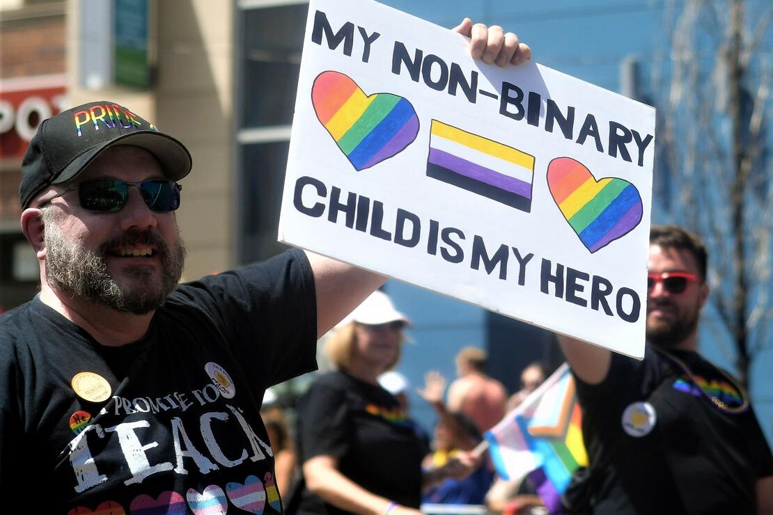 En voksen mann med regnbuecaps holder en plakat hvor det står "my non-binary child is my hero" med regnbuehjerter og ikke-binær-flagget. 