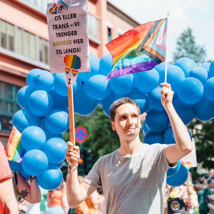 Fra Oslo Pride 2023. Person som holder et progress pride-flagg og plakat hvor det står "cis eller trans - vi trenger bønder her til lands!"