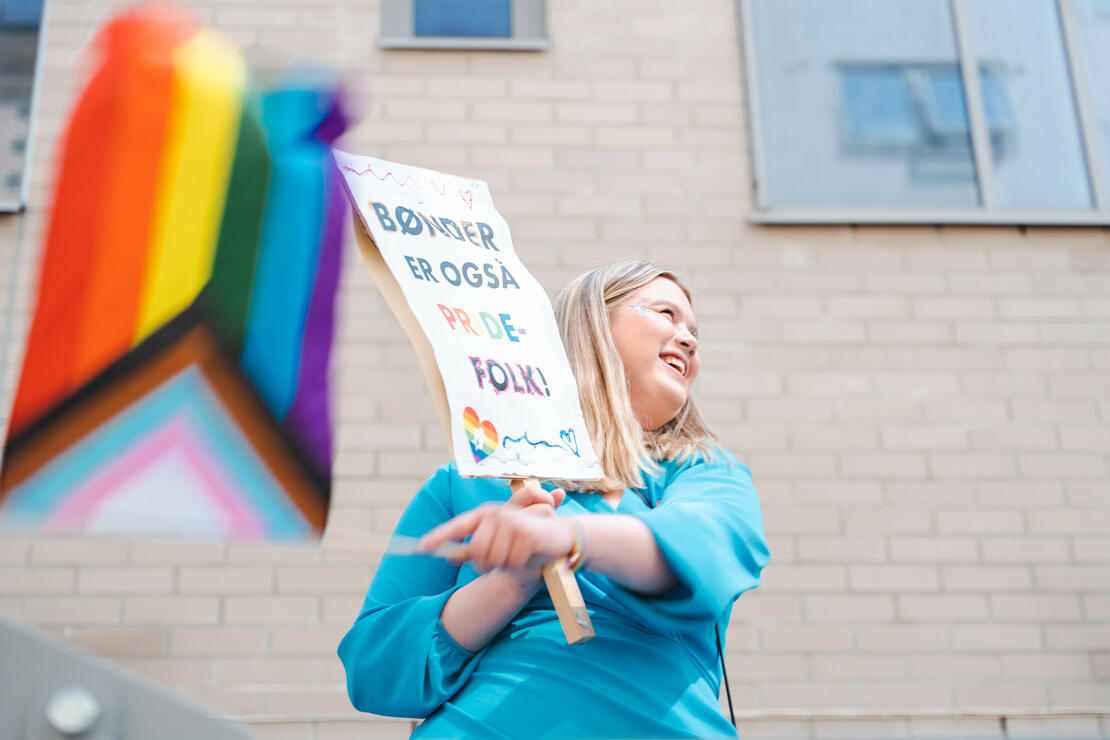 Oslo Pride 2023. Person som smiler bredt, veiver med progress pride-flagg og holder plakat hvor det står "bønder er også pride-folk!"